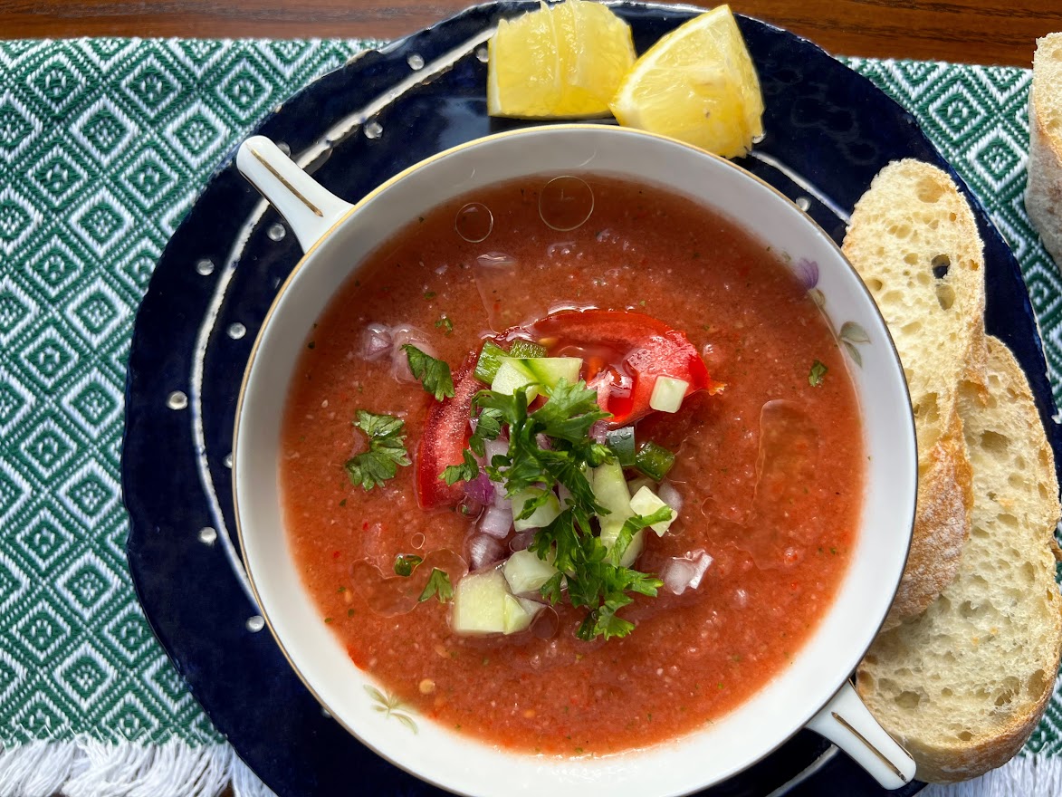 Photo of bowl of Gazpacho Andaluz on blue plate with lemon wedges and sliced baguette