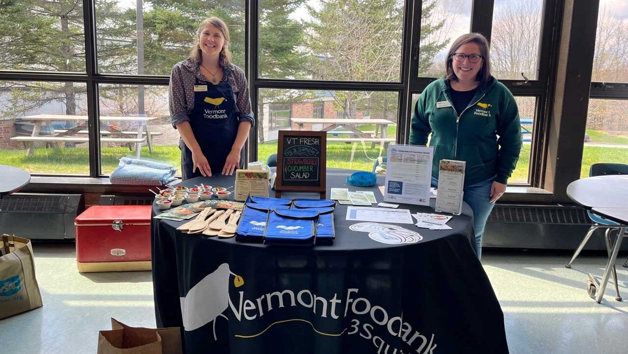 Photo of two Vermont Foodbank 3SquaresVT team members looking at the camera and standing behind a table with informational materials on it.